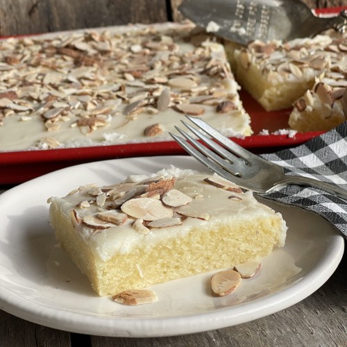 Slice of White Almond Texas Sheet cake on a plate with red serving pan of cake in the back ground